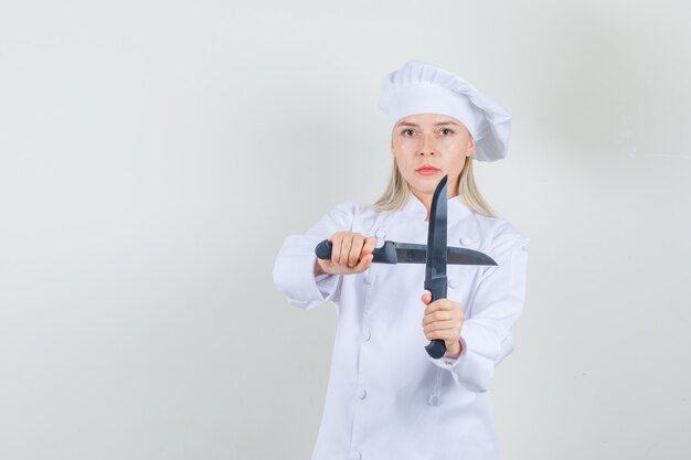 Chef mujer sosteniendo cuchillos en uniforme blanco y mirando serio