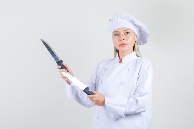 Chef mujer sosteniendo cuchillos en uniforme blanco y mirando serio