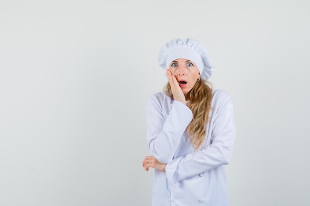 Chef mujer sosteniendo la mano en la mejilla en uniforme blanco y mirando sorprendido