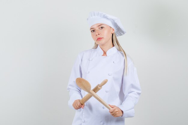 Chef mujer sosteniendo un rodillo y una cuchara de madera en uniforme blanco y mirando serio