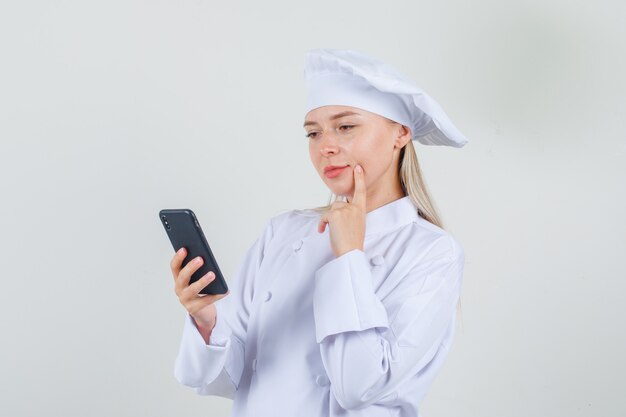 Chef mujer sosteniendo smartphone con el dedo en la mejilla en uniforme blanco.