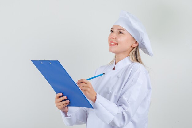 Chef mujer tomando notas en el portapapeles en uniforme blanco y mirando alegre