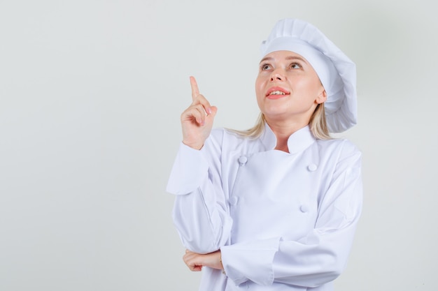 Chef mujer en uniforme blanco apuntando con el dedo hacia arriba y mirando alegre