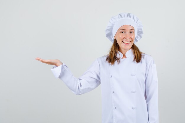 Chef mujer en uniforme blanco dando la bienvenida o mostrando algo con la palma abierta y mirando contento, vista frontal.