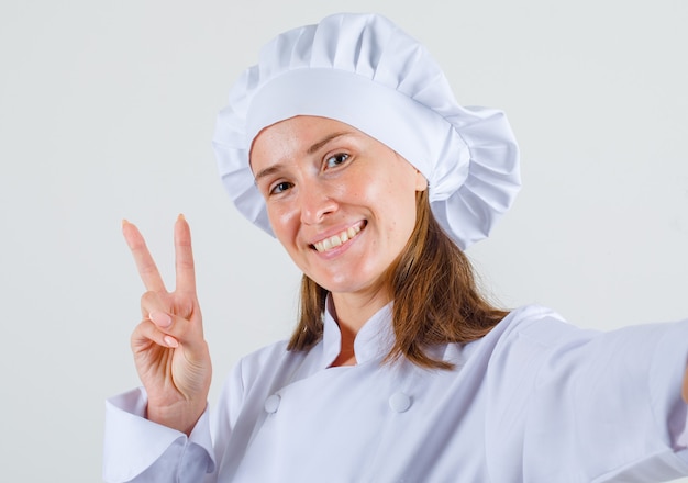 Chef mujer en uniforme blanco mostrando gesto de paz y mirando contento