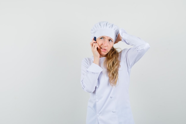 Chef mujer en uniforme blanco rascándose la cabeza mientras habla por teléfono celular y parece vacilante