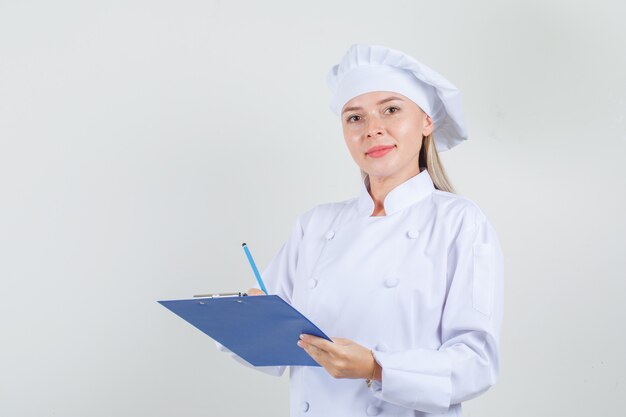 Chef mujer en uniforme blanco sosteniendo lápiz y portapapeles y mirando alegre