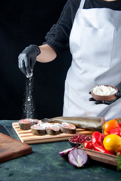 Chef mujer vista frontal con delantal que cubre rebanadas de pescado crudo con verduras frescas de harina en tablero de madera cuchillo de tazón de harina en la mesa de la cocina