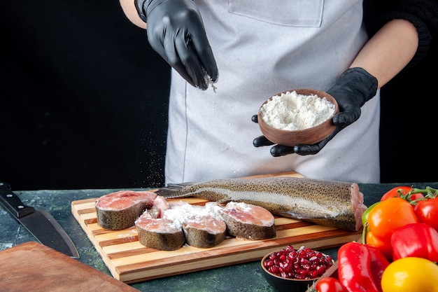 Chef mujer vista frontal con delantal que cubre rodajas de pescado crudo con verduras frescas de harina en un tazón de harina de tablero de madera en la mesa de la cocina