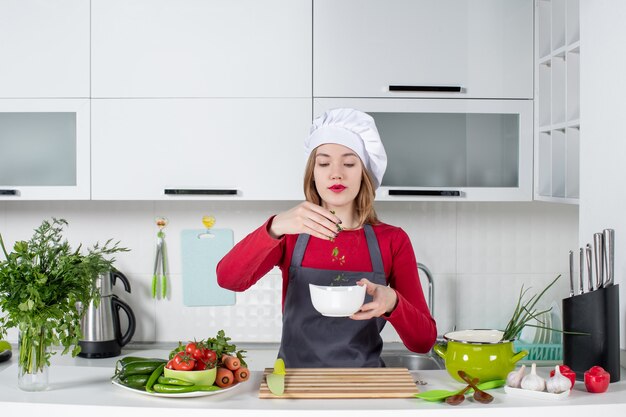 Chef mujer vista frontal en uniforme de pie detrás de la mesa de la cocina sosteniendo un tazón con verduras