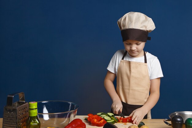 Chef de niño varón enfocado en delantal y sombrero cortando verduras para lasaña vegetariana