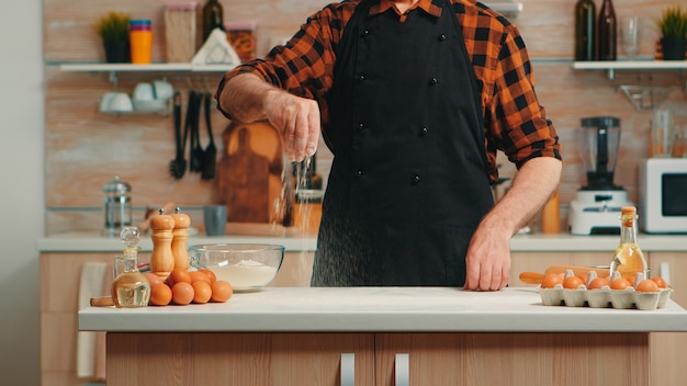 Foto gratuita chef panadero experimentado que usa harina de trigo para esparcirla para la preparación de alimentos. hombre senior jubilado con bonete y delantal espolvorear tamizado tamizar ingredientes a mano para hornear pan y pizza casera