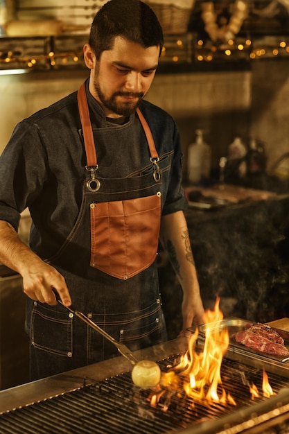 Chef profesional cocinando verduras de cebolla y carne a la parrilla con fuego y humo hermoso hombre concentrado en la preparación de los antecedentes alimentarios de la cocina moderna del restaurante