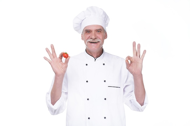 Chef profesional de edad avanzada en uniforme blanco y sombrero, sosteniendo tomate mientras permanece en la pared blanca