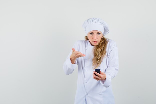 Chef de sexo femenino que mira el teléfono celular en uniforme blanco y parece confundido.