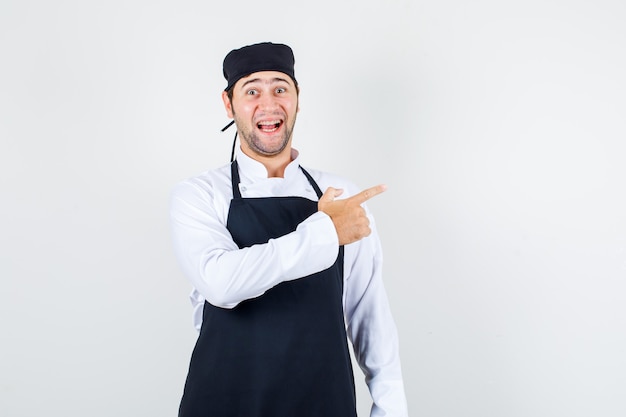 Chef de sexo masculino apuntando al lado en uniforme, delantal y mirando alegre, vista frontal.