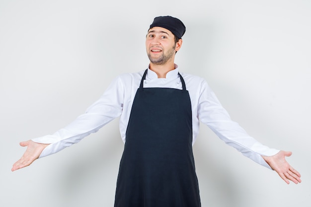 Chef de sexo masculino extendiendo los brazos en uniforme, delantal y mirando alegre, vista frontal.