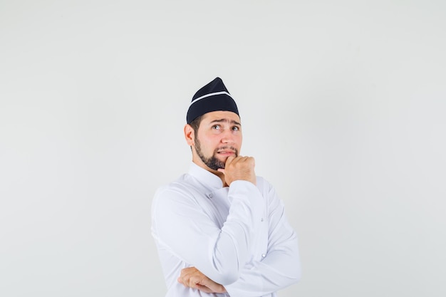 Chef de sexo masculino mirando hacia arriba con la mano en la barbilla en uniforme blanco y mirando pensativo. vista frontal.