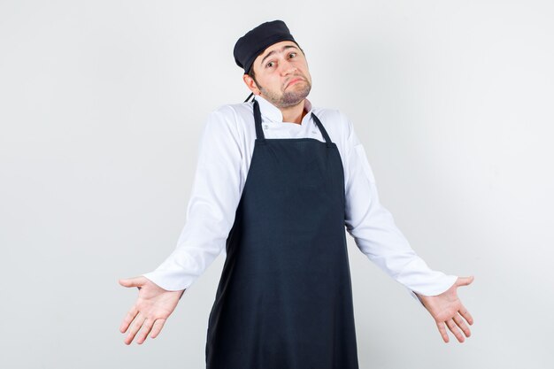 Chef de sexo masculino que muestra un gesto de impotencia en uniforme, delantal y mirando confundido, vista frontal.