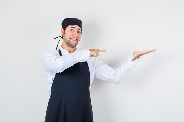 Chef de sexo masculino que muestra la palma extendiéndose a un lado en uniforme, delantal y con aspecto alegre. vista frontal.