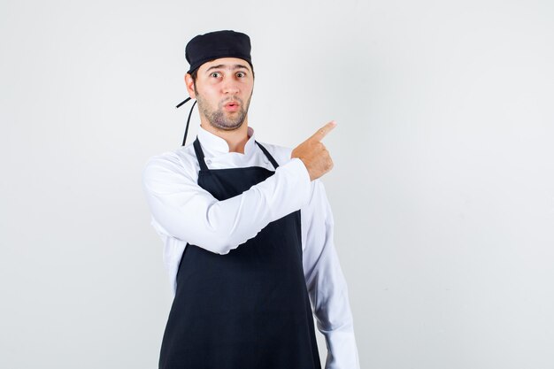 Chef de sexo masculino en uniforme, delantal apuntando con el dedo a un lado y mirando sorprendido, vista frontal.