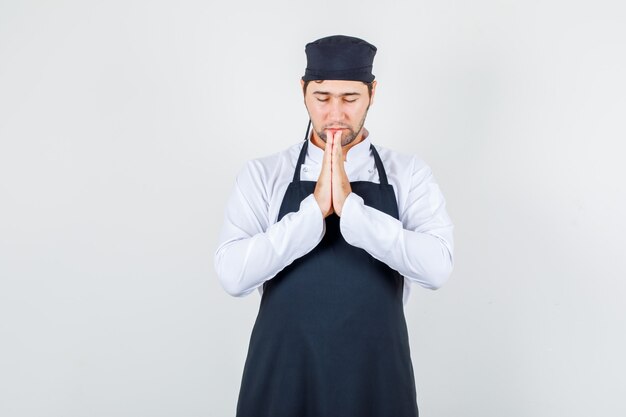 Chef de sexo masculino en uniforme, delantal cogidos de la mano en gesto de oración y mirando en silencio, vista frontal.