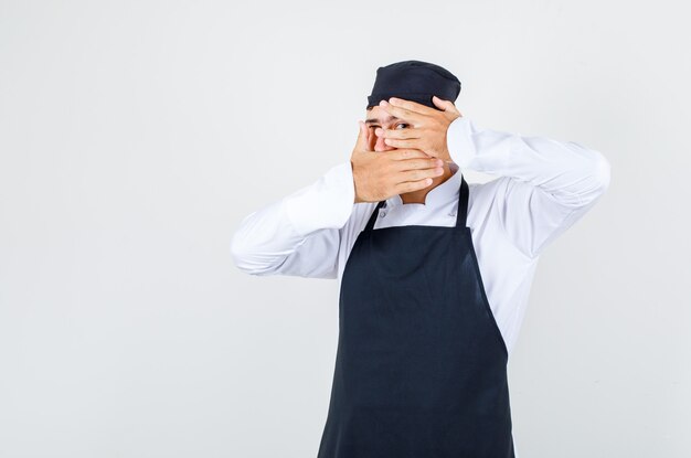 Foto gratuita chef de sexo masculino en uniforme, delantal que cubre la cara y mirando a través de los dedos, vista frontal.