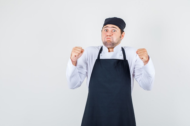 Chef de sexo masculino en uniforme, delantal soplando las mejillas con los puños cerrados, vista frontal.