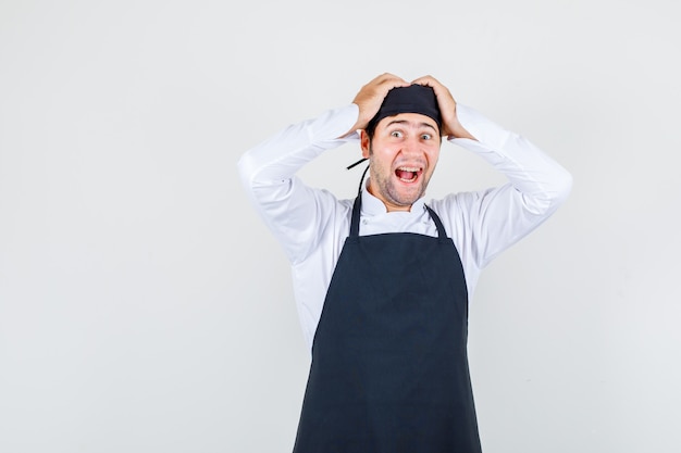 Chef de sexo masculino en uniforme, delantal sosteniendo la cabeza con las manos mientras grita y parece emocionado, vista frontal.