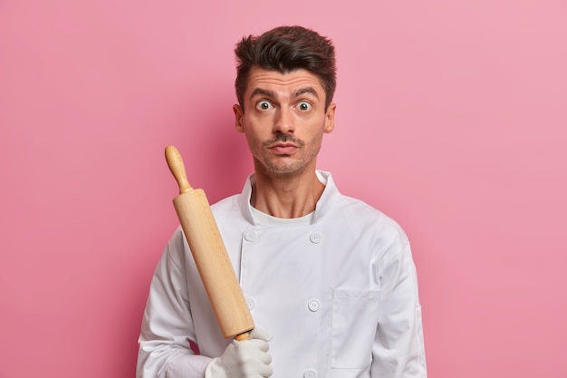 Chef sorprendido con herramienta de cocina, vestido con uniforme blanco, panadero ocupado sostiene un rodillo