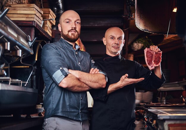 Chef sosteniendo un bistec fresco y su asistente de pie cerca con los brazos cruzados en la cocina de un restaurante.