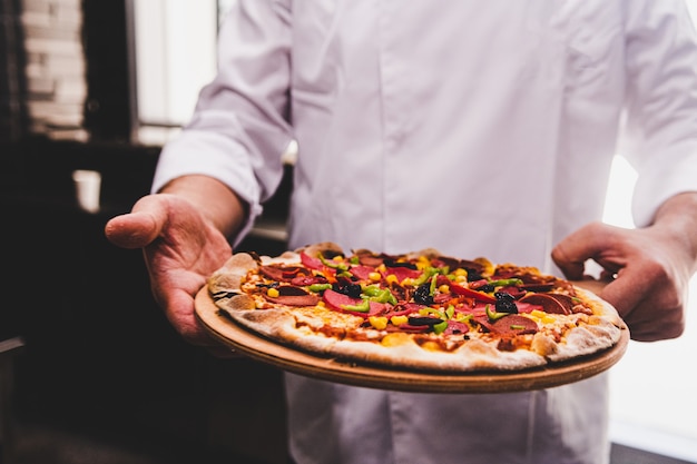 Chef sosteniendo un plato de madera con una deliciosa pizza en la cocina