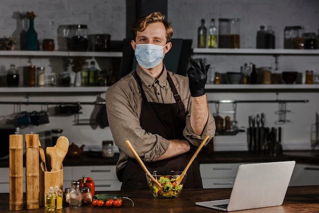 Chef de tiro medio con máscara mezclando ingredientes de ensalada cerca de la computadora portátil