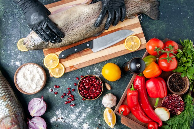 Chef de vista superior con guantes negros sosteniendo pescado crudo en tablero de madera molinillo de pimienta harina tazón de granada semillas en un tazón en la mesa