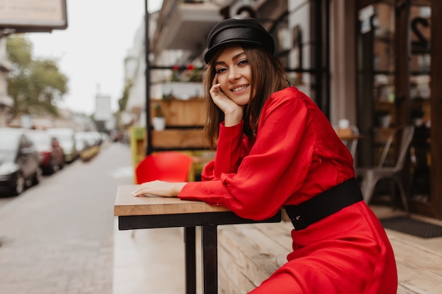 Chica de 23 años de Francia posando sentada en un café de la calle. Tiro acogedor de dama elegante en vestido rojo con mangas anchas