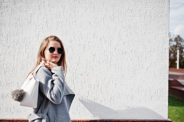 Chica en abrigo gris con gafas de sol y bolso de mano contra la pared blanca