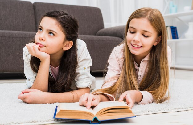 Chica aburrida acostada con su amiga leyendo el libro en la sala de estar
