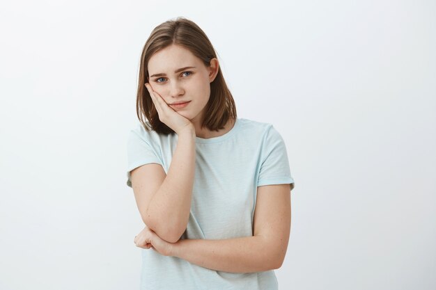 Chica aburrida de la rutina. Retrato de mujer europea tranquila y guapa que se inclina la cabeza en la palma mirando con mirada cansina e indiferente sin nada que hacer de pie contra la pared gris