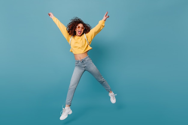 Chica activa muestra lengua. Mujer en suéter amarillo y jeans saltando felizmente en espacio azul aislado.