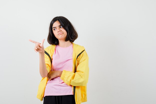 Chica adolescente apuntando hacia arriba con el dedo en chándal amarillo, camiseta y mirando sorprendido, vista frontal.