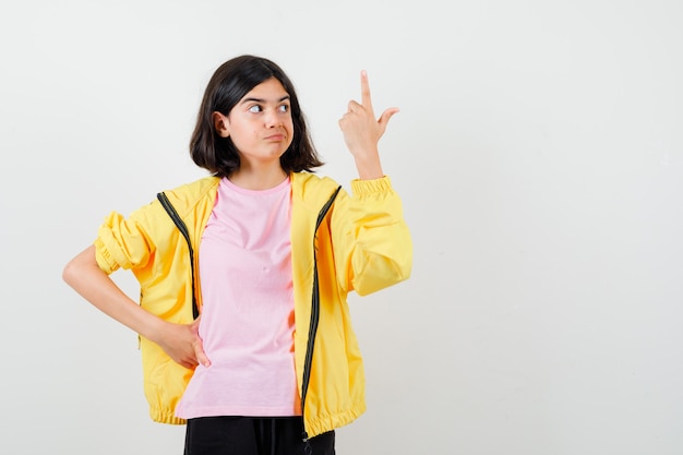Chica adolescente apuntando hacia arriba, sosteniendo la mano en la cintura en chándal amarillo, camiseta y mirando disgustado, vista frontal.