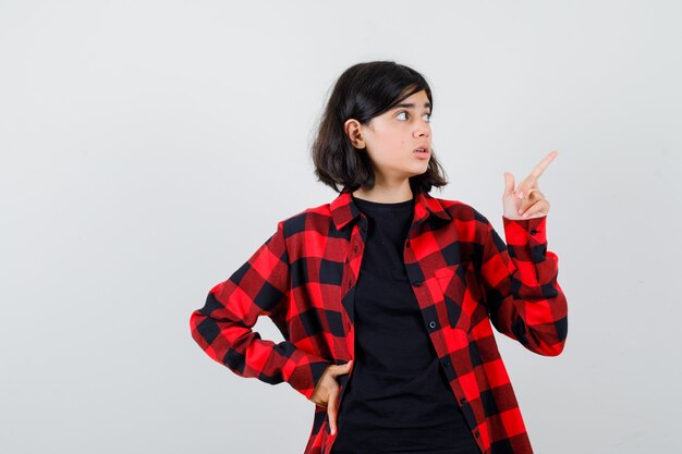 Chica adolescente apuntando a la esquina superior derecha en camiseta, camisa a cuadros y mirando perplejo, vista frontal.