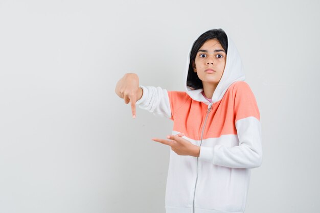 Chica adolescente apuntando hacia un lado y hacia abajo en chaqueta blanca y mirando atenta.