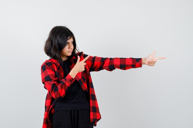 Chica adolescente apuntando a un lado en camiseta, camisa a cuadros y mirando enfocado, vista frontal.