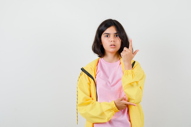 Chica adolescente apuntando hacia el lado derecho y hacia arriba en camiseta, chaqueta y mirando sorprendido. vista frontal.