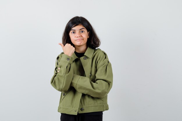 Chica adolescente apuntando hacia el lado izquierdo con el pulgar en camiseta, chaqueta verde y mirando con cuidado. vista frontal.
