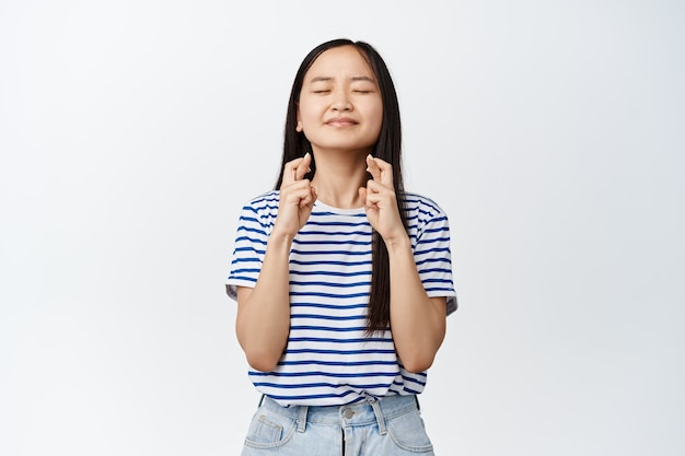 Chica adolescente asiática esperanzada que pide deseos, cierra los ojos y cruza los dedos para la buena suerte, reza, anticipa que algo suceda, de pie en blanco.