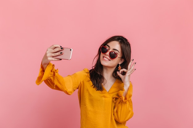 Chica adolescente en blusa brillante y gafas de sol inusuales muestra el signo "OK", haciendo selfie.