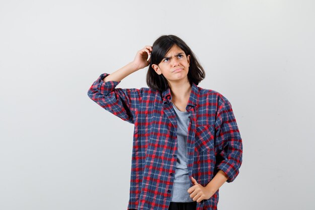 Chica adolescente en camisa a cuadros rascándose la cabeza y mirando perplejo, vista frontal.