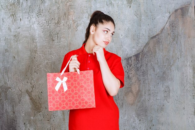 Chica adolescente en camisa roja sosteniendo una bolsa roja y parece confundida y pensativa.